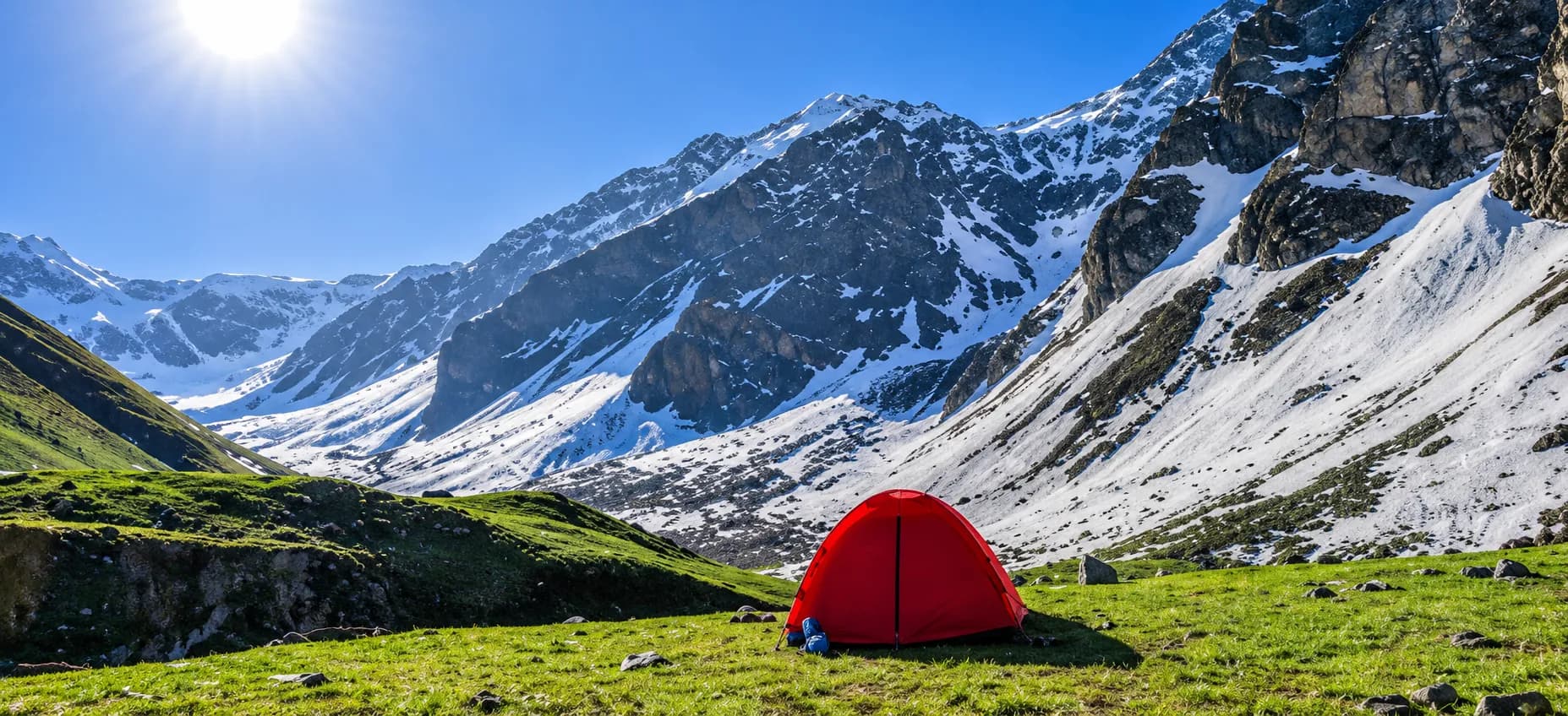 A red tent camp on a green meadow below snow-covered peaks, typical of the Buran Ghati trek