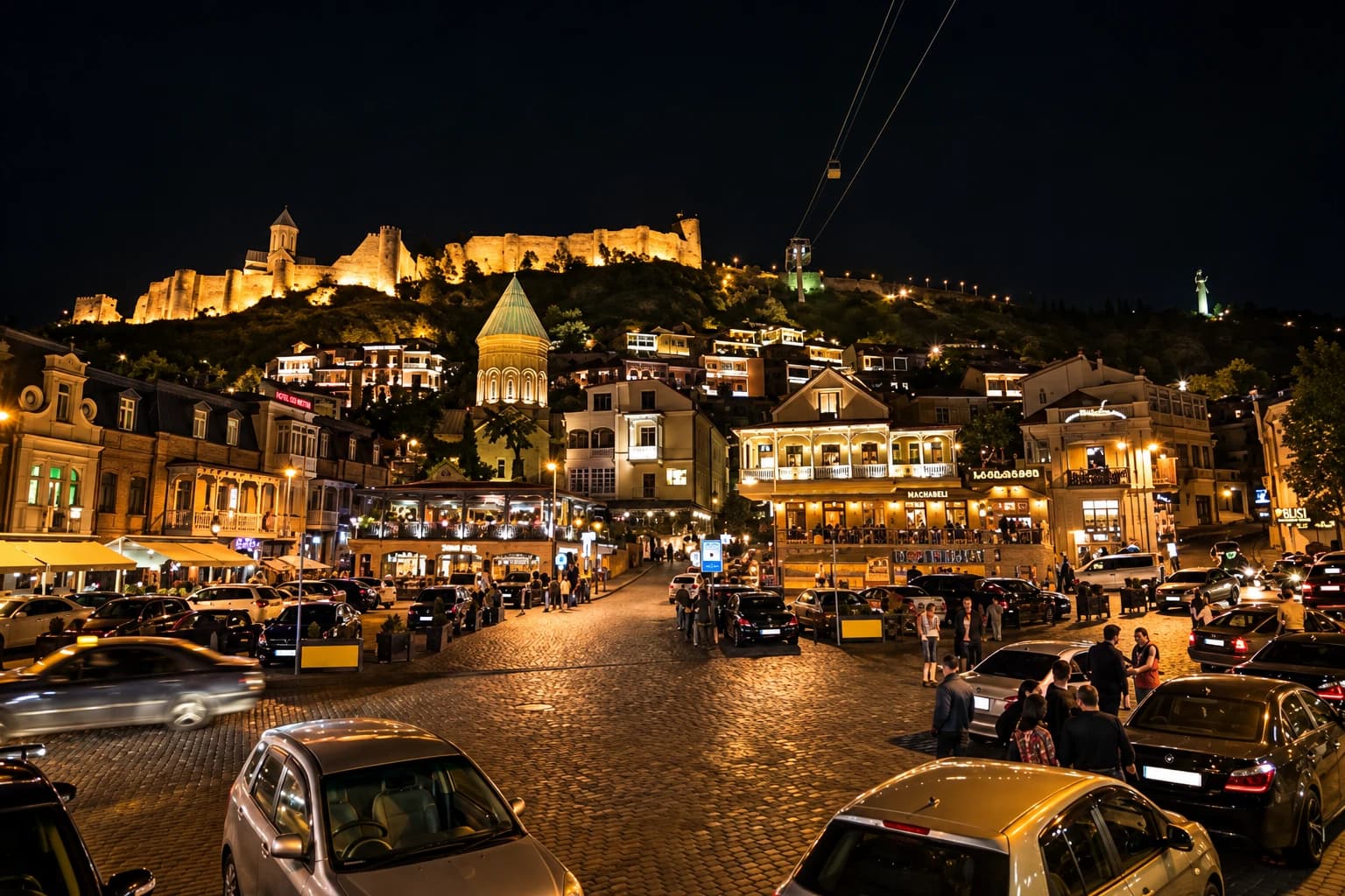 Tbilisi's Old Town lit up at night with the illuminated Narikala Fortress on the hill above and a lively cobblestone street below